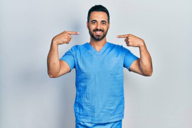 Sakallı yakışıklı, mavi erkek hemşire üniforması giyen neşeli gülümseyen ve parmaklarıyla ve ağzıyla işaret eden bir İspanyol. diş sağlığı kavramı. 