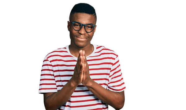 若いアフリカ系アメリカ人男性を着てカジュアルなTシャツと眼鏡を手で祈る一緒に笑顔自信を許しを求めて — ストック写真