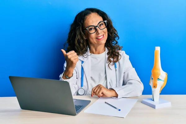 Beautiful middle age woman doctor at orthopedic clinic smiling with happy face looking and pointing to the side with thumb up.