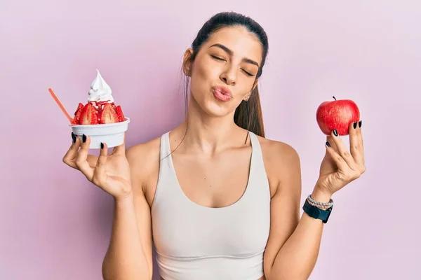 穿着运动服 拿着苹果和冰淇淋的年轻的惊慌失措的女人看着镜头 飞吻着一个可爱而性感的吻 爱的表达 — 图库照片