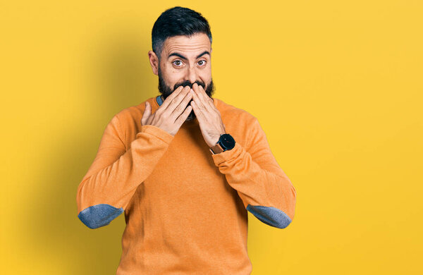 Латиноамериканец с бородой в обычном зимнем свитере смеется и смущенно хихикает, прикрывая рот руками, сплетнями и скандалом 