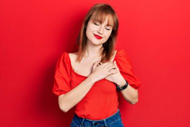 Kırmızı tişörtlü kızıl saçlı genç kadın elleri göğsünde, gözleri kapalı ve yüzünde minnet dolu bir jest ile gülümsüyor. sağlık kavramı. 