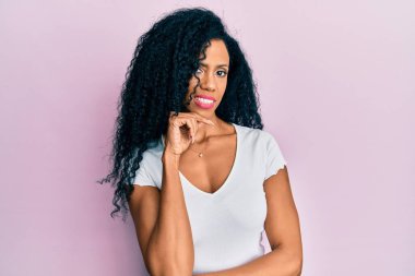 Orta yaşlı, beyaz tişört giyen Afro-Amerikalı bir kadın kameraya kollarını kavuşturup çenesine dokunarak gülümsüyor. Olumlu düşünmek. 