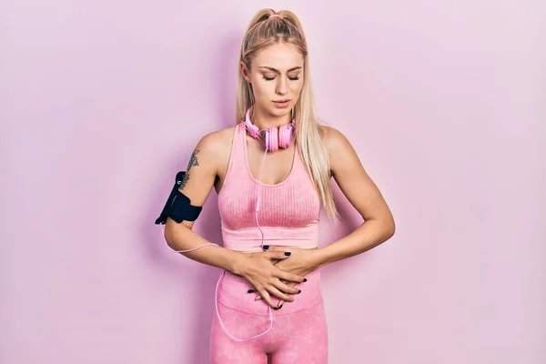 Young beautiful caucasian woman wearing gym clothes and using headphones with hand on stomach because indigestion, painful illness feeling unwell. ache concept.