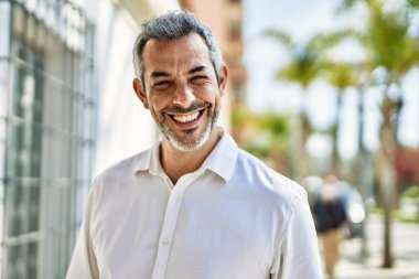 Orta yaşlı, kır saçlı, gülümseyen bir adam şehirde mutlu bir şekilde duruyor..