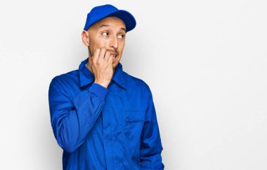 Kel, sakallı, işçi tulumu giyen, stresli ve elleri ağız yiyen tırnakları olan bir adam. anksiyete. 