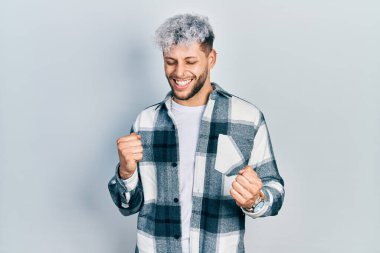 Modern boyalı saçlı, mutlu ve heyecanlı genç İspanyol adam kolları havada, gülümseyen ve başarı için bağıran kazanan jestler yapıyor. kutlama konsepti. 