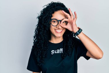 Kıvırcık saçlı, personel tişörtü giyen, el hareketleriyle gülümseyen, mutlu yüzüyle parmaklarının arasından bakan İspanyol bir kadın.. 