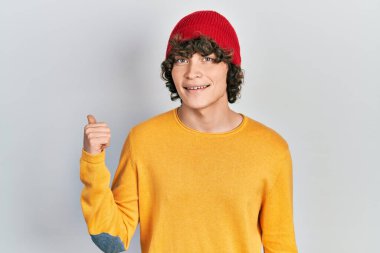 Yakışıklı, yün şapkalı, mutlu yüzlü, gülümseyen ve baş parmağıyla yan tarafı gösteren genç bir adam.. 