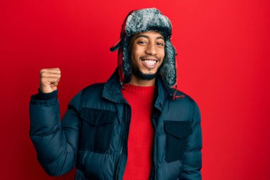 Sakallı, kışlık şapkalı ve ceketli gülümseyen, gülümseyen ve baş parmağıyla yüzünü gösteren Afro-Amerikan genç adam.. 