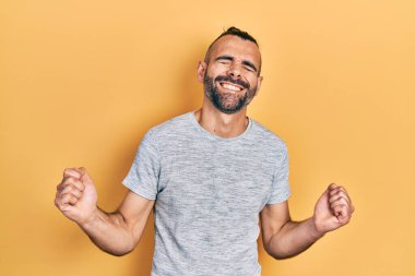 Gündelik kıyafetler giyen genç İspanyol adam çok mutlu ve heyecanlı. Kazanan jestler yapıyor. Kolları havada, gülümsüyor ve başarı için bağırıyor. kutlama konsepti. 