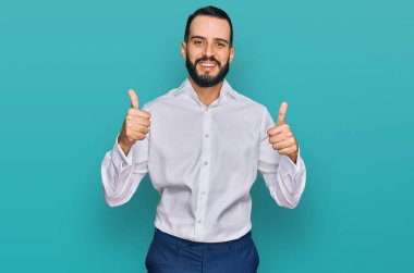 Sakallı, iş gömleği giyen, başarı işareti takan, el hareketi yapan, gülümseyen ve mutlu genç bir adam. Neşeli ifade ve kazanan jest. 