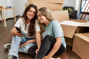 Genç çift yeni evinde akıllı telefon kullanarak mutlu bir şekilde gülümsüyor..