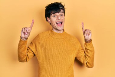 Yakışıklı hippi genç adam gündelik sarı süveter giyiyor şaşkın şaşkın gülüyor, parmaklarıyla işaret ediyor ve kollarını kaldırıyor.. 