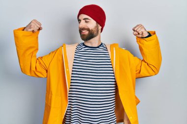 Sakallı, sarı yağmurluk giyen beyaz adam gururla gülümseyen kol kaslarını gösteriyor. fitness konsepti. 