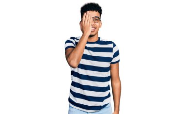 Genç Afrikalı Amerikalı bir adam günlük kıyafetler ve gözlük takıyor. Bir gözünü eliyle kapatıyor. Yüzünde kendine güvenen bir gülümseme ve sürpriz bir duygu var.. 