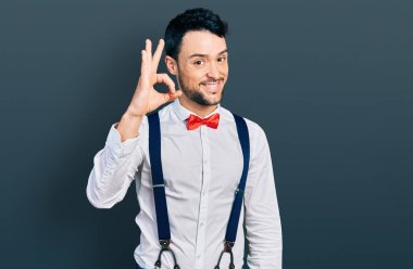 Sakallı, hippi kılıklı, papyonlu, pantolon askısı takan, gülümseyen ve el ve parmaklarıyla imza atan İspanyol bir adam. başarılı ifade. 