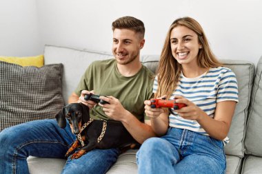 Genç İspanyol çift gülümseyerek evde köpekle kanepede oturup video oyunu oynuyor..