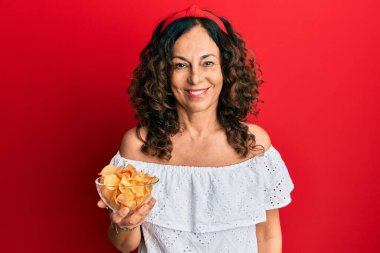 Orta yaşlı İspanyol kadın elinde patates cipsi tutarken pozitif ve mutlu görünüyor. Kendine güvenen bir gülümsemeyle ayakta duruyor ve dişlerini gösteriyor. 