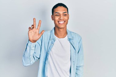 Genç, Afro-Amerikalı, günlük giysiler giymiş, üç numaralı parmaklarıyla işaret ederken, kendinden emin ve mutlu bir şekilde gülümsüyor.. 