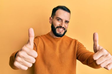 Sakallı genç adam sıradan bir kazak giyiyor. Elleriyle olumlu bir jest yapıyor. Gülümsüyor ve başarı için seviniyor. kazanan jest. 