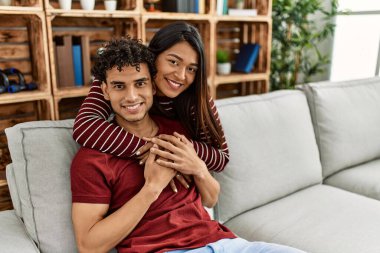 Mutlu gülümseyen ve sarılan genç Latin çift evdeki kanepede oturuyor..