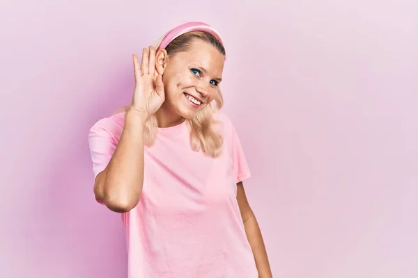 耳を傾け 噂やゴシップに耳を傾け手で笑みを浮かべてカジュアルなピンクのTシャツを着て美しい白人ブロンドの女性 聴覚障害の概念 — ストック写真
