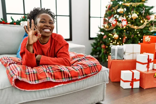 若いアフリカ系アメリカ人女性がクリスマスツリーでソファの上に横たわり 手と指でOkサインをして積極的に笑っています 成功した表現 — ストック写真