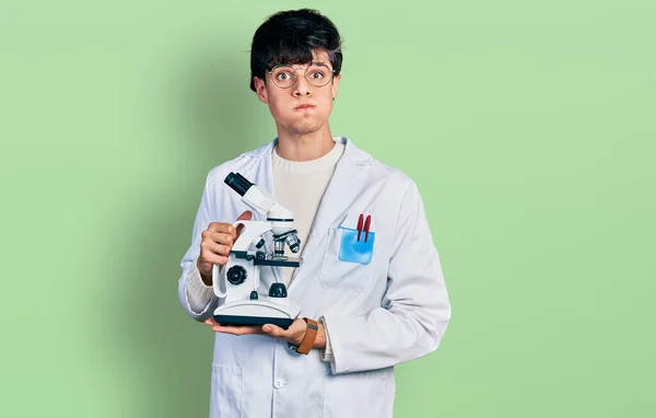 Joven Hipster Guapo Con Bata Laboratorio Sosteniendo Microscopio Soplando Mejillas —  Fotos de Stock