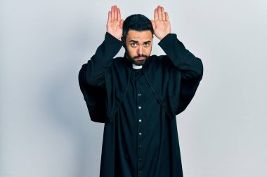 Sakallı, Katolik rahip cübbesi giyen, tavşan kulağı hareketi yapan, elleri avuçlarında, alaycı ve şüpheci görünen yakışıklı bir İspanyol. Paskalya tavşanı konsepti. 