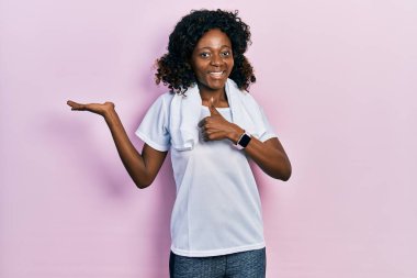 Afro-Amerikalı genç bir kadın spor kıyafetleri ve havlu giyiyor avuç içini gösteriyor ve baş parmaklarıyla iyi bir hareket yapıyor, mutlu ve neşeli gülümsüyor. 