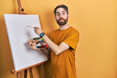 Beyaz, sakallı bir adam boyacı sehpasında paletle resim çizerken kameraya bakıyor ve öpüşürken çok güzel ve seksi oluyor. aşk ifadesi. 