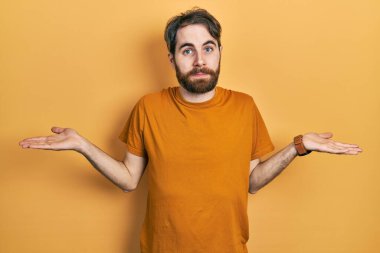 Sakallı, sarı tişörtlü beyaz bir adam her iki elinde avuçlarını açmış gülümsüyor, sunuyor ve reklam karşılaştırması yapıyor ve dengede duruyor. 