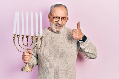 Sakallı yakışıklı son sınıf öğrencisi elinde menorah Hanuka Musevi mumu mutlu ve pozitif gülümsüyor, başparmağı mükemmel ve onay işareti yapıyor. 