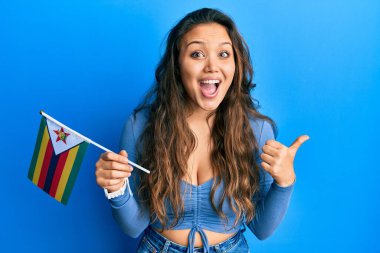 Genç İspanyol kız elinde zimbabwe bayrağı, baş parmağını kaldırmış, ağzı açık gülümsüyor. 