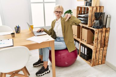 Ofiste çalışan beyaz, güzel bir iş kadını kızgın yüzüyle pilates topunda oturuyor. Negatif işaretler baş parmakları aşağıda, reddedilme konsepti ise reddedilmiş. 