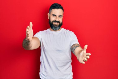 Latin asıllı genç bir adam, günlük beyaz tişört giyip kameraya bakıyor ve sarılmak için kollarını açmış gülümsüyor. Mutluluğu kucaklayan neşeli bir ifade.. 