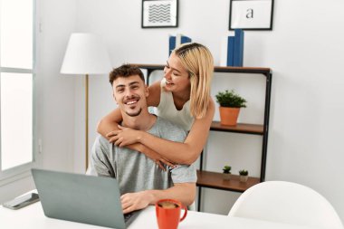 Genç, beyaz bir çift kucak açıp dizüstü bilgisayar kullanıyor. Evdeki masanın üstünde oturuyorlar..