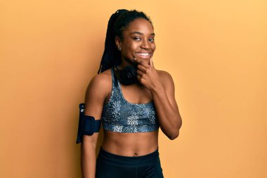 Saçları örgülü, spor kıyafetleri ve kol bandı takmış, kolları çapraz ve elleri çenesinde gülümseyerek kameraya bakan Afro-Amerikalı bir kadın. Olumlu düşünmek. 