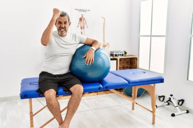 Orta yaşlı İspanyol bir adam acı dindirme kliniğinde elinde pilates topu tutarken kızgın ve kızgın yumruk kaldırırken öfkeyle bağırıyor. öfke ve agresif konsept. 