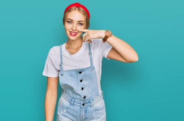 Kafkasyalı genç bir kız, günlük kıyafetler giyip, el ve burnu parmakla göstererek, neşeli bir şekilde gülümsüyor. güzellik konsepti 