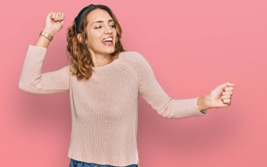 Güzel, beyaz bir kadın gündelik bir süveter giyiyor mutlu ve neşeli dans ediyor, gülümsüyor rahat ve kendinden emin bir şekilde müzik dinliyor. 