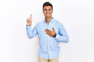 İş gömleği giyen genç İspanyol adam izole edilmiş arka planda durup gülümsüyor, elleri göğsünde ve parmakları havada küfür ediyor, sadakat yemini ediyor. 