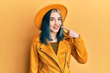 Sarı şapkalı ve deri ceketli genç modern bir kız, el parmağı ve burnu işaret ederek, neşeli bir şekilde gülümsüyor. güzellik konsepti 