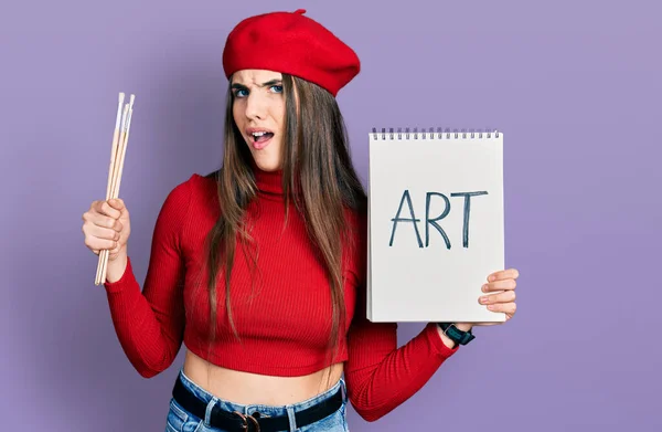Young Brunette Teenager Holding Art Notebook Paintbrushes Clueless Confused Expression — Stock Photo, Image