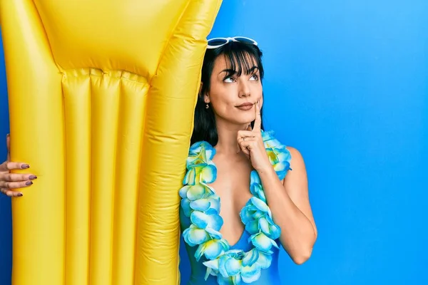 Young brunette woman with bangs wearing swimsuit and holding summer float serious face thinking about question with hand on chin, thoughtful about confusing idea