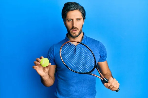 Junger Hispanischer Tennisspieler Mit Schläger Und Ball Schockgesicht Sieht Skeptisch — Stockfoto