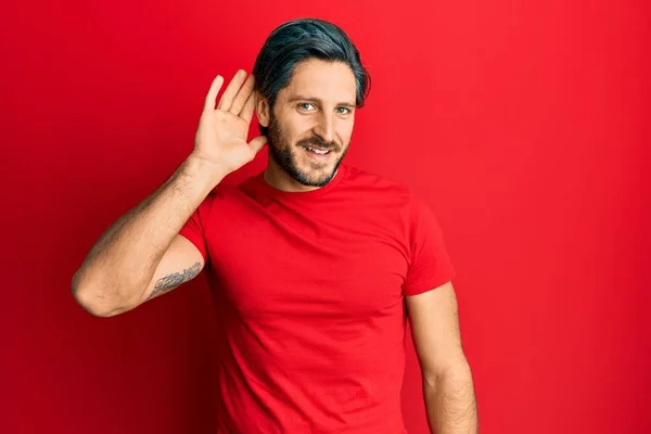噂やゴシップに耳を傾ける耳の上に手で笑みを浮かべてカジュアルな赤いTシャツを着て若いヒスパニック系の男 聴覚障害の概念 — ストック写真