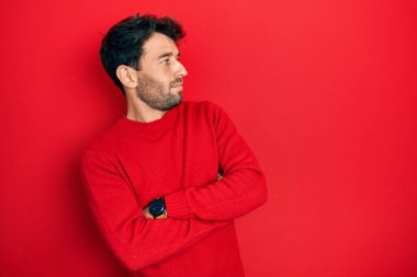 Sakallı, sıradan kırmızı süveter giyen, kolları çapraz ve kendinden emin yakışıklı bir adam. 