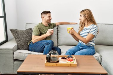 Genç İspanyol çift mutlu bir şekilde gülümsüyor. Evdeki kanepede oturup kahvaltı yapıyorlar..